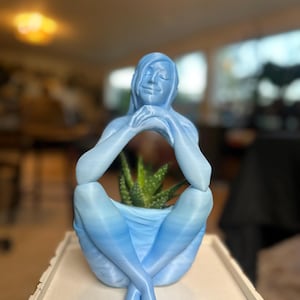 May include: A light blue, 3D-printed planter shaped like a person in a seated yoga pose. The figure's arms are crossed, and the legs are crossed at the ankles. A small succulent plant is inside the planter. The figure is on a white table.