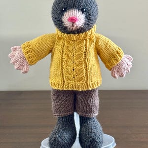 May include: A knitted mole doll with a grey head, pink nose, and a yellow cable-knit sweater. The doll wears brown shorts, grey boots, and has pink hands. It stands on a clear plastic base.