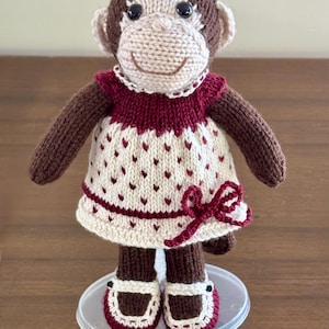 May include: A hand-knitted monkey doll with dark brown fur and a cream-coloured face. The monkey wears a cream-coloured dress featuring a small red heart pattern, a dark red collar, a matching red bow at the hem, and dark red and cream shoes.