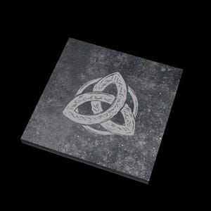 May include: A square, dark gray stone coaster with a Celtic knot design etched in light gray. The knot is a stylized, three-part design, with each part interlocked. The coaster has a speckled texture and a slightly beveled edge.