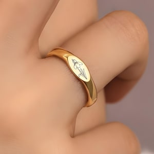 Puede incluir: Un anillo dorado con una cara ovalada con un diseño floral negro. El anillo se lleva en un dedo, mostrando su superficie pulida y reflectante. El diseño del anillo es simple y elegante.