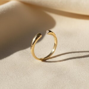 May include: A gold-colored open ring with a small, clear gemstone set at one end. The ring has a smooth, polished finish and a modern, minimalist design. The ring is displayed on a neutral-toned fabric, highlighting its elegant simplicity.