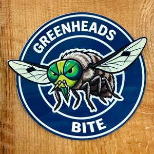 May include: A round sticker with a blue background and white border. The sticker features a cartoon illustration of a green-eyed fly with the text "GREENHEADS BITE" in white. The fly has black and white wings and a green and yellow head.