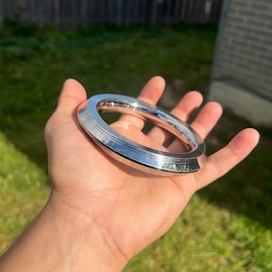May include: A silver-colored, thick bangle bracelet with a rounded, ridged design. The bracelet is held in a hand against a background of green grass and a wooden fence.