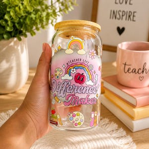 Könnte beinhalten: Ein klares Glas mit einem Holzdeckel, verziert mit bunten Grafiken und den Worten "Difference Maker". Das Design umfasst einen Regenbogen, einen Apfel und andere Illustrationen zum Thema Schule. Das Glas wird von einer Hand gehalten.