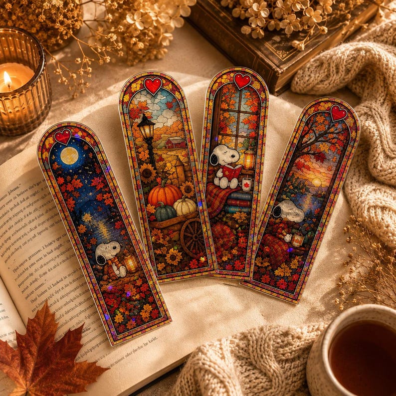 Four stained-glass-style autumn bookmarks depicting Snoopy scenes laid out on an open book.