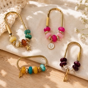 May include: Four gold-toned metal U-shaped keychains featuring colorful beads and small dangling letter charms. One keychain has a white circular charm with the letter U, while the others feature gold metal letter charms K, P, and R.
