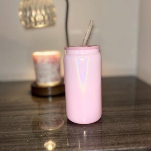 K&ouml;nnte beinhalten: Ein rosa, schillernder Becher mit einem durchsichtigen Strohhalm. Der Becher ist zylindrisch mit einem Deckel und steht auf einer dunklen, gl&auml;nzenden Oberfl&auml;che. Eine dekorative Lampe und eine Kerze sind im Hintergrund.