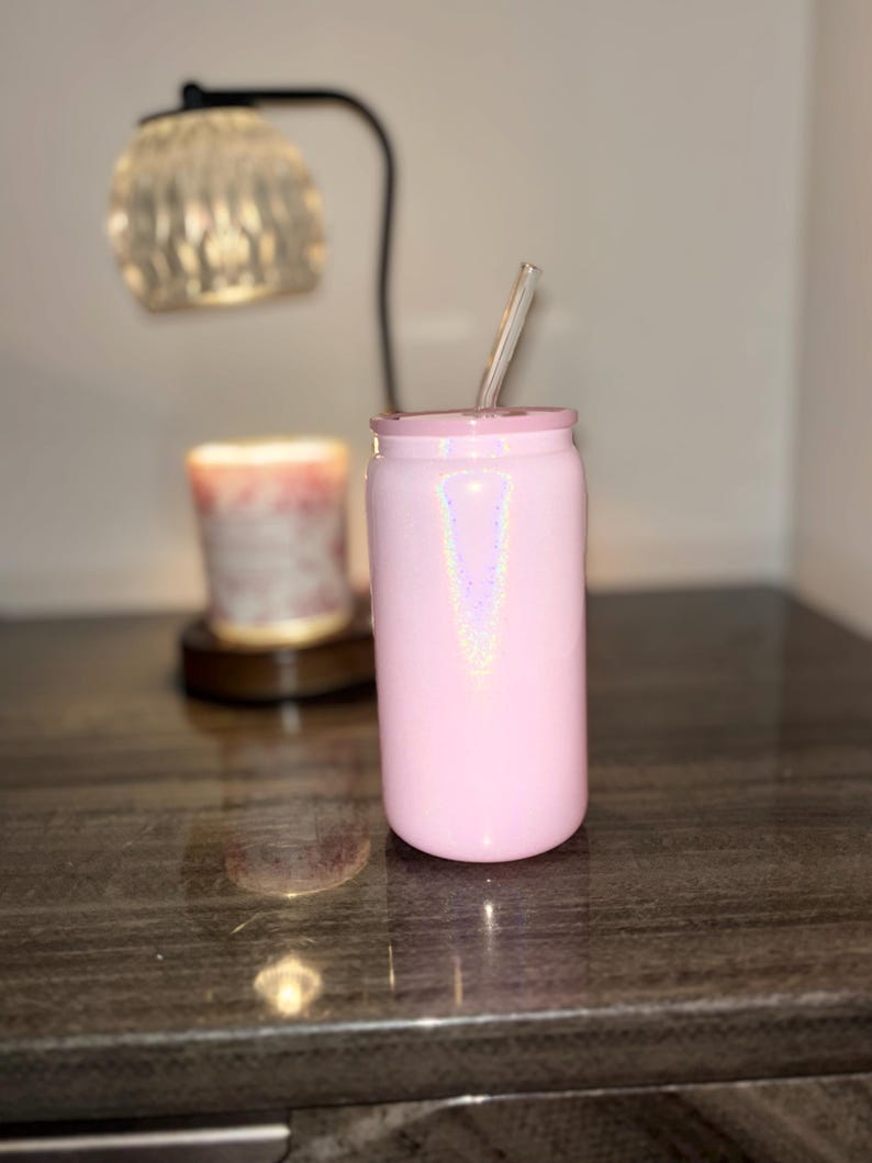 K&ouml;nnte beinhalten: Ein rosa, schillernder Becher mit einem durchsichtigen Strohhalm. Der Becher ist zylindrisch mit einem Deckel und steht auf einer dunklen, gl&auml;nzenden Oberfl&auml;che. Eine dekorative Lampe und eine Kerze sind im Hintergrund.