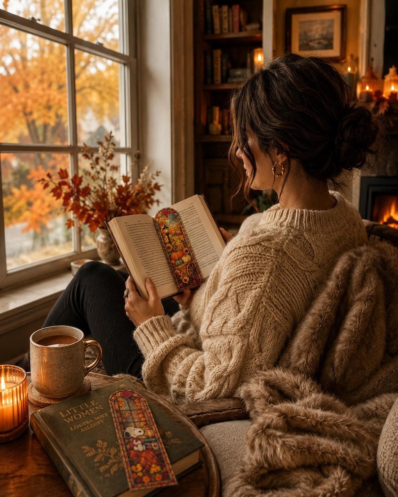 A woman in a cream sweater reads a book by a window next to a mug and another book with a stained-glass bookmark.