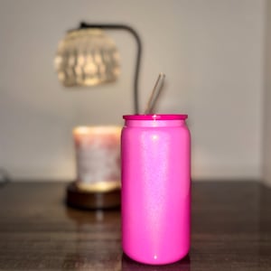 K&ouml;nnte beinhalten: Ein leuchtend pinkfarbener, mit Glitzer &uuml;berzogener Becher mit passendem pinkfarbenem Rand und einem durchsichtigen Strohhalm. Das zylindrische Trinkgef&auml;&szlig; ist so gestaltet, dass es einer Limonadendose &auml;hnelt. Der Hintergrund zeigt eine verschwommene Lampe und Kerze auf dunkler Oberfl&auml;che.