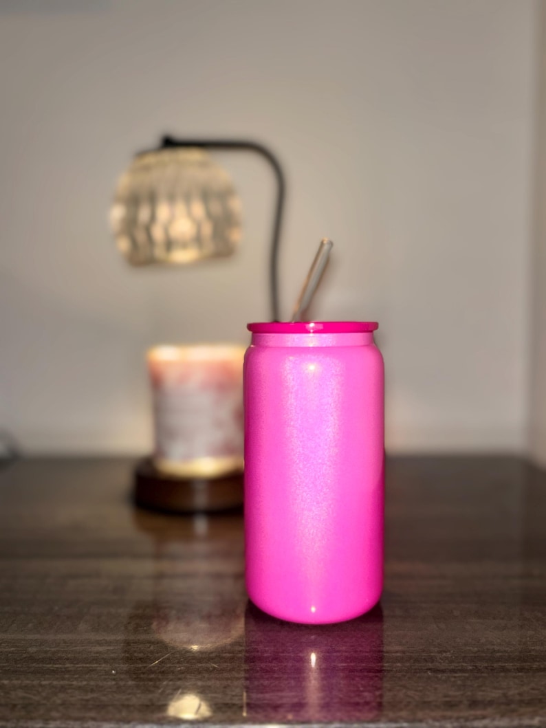 K&ouml;nnte beinhalten: Ein leuchtend pinkfarbener, mit Glitzer &uuml;berzogener Becher mit passendem pinkfarbenem Rand und einem durchsichtigen Strohhalm. Das zylindrische Trinkgef&auml;&szlig; ist so gestaltet, dass es einer Limonadendose &auml;hnelt. Der Hintergrund zeigt eine verschwommene Lampe und Kerze auf dunkler Oberfl&auml;che.