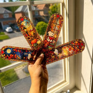 A hand holds up four fan-arranged holographic bookmarks with fall-themed stained glass designs against a window.