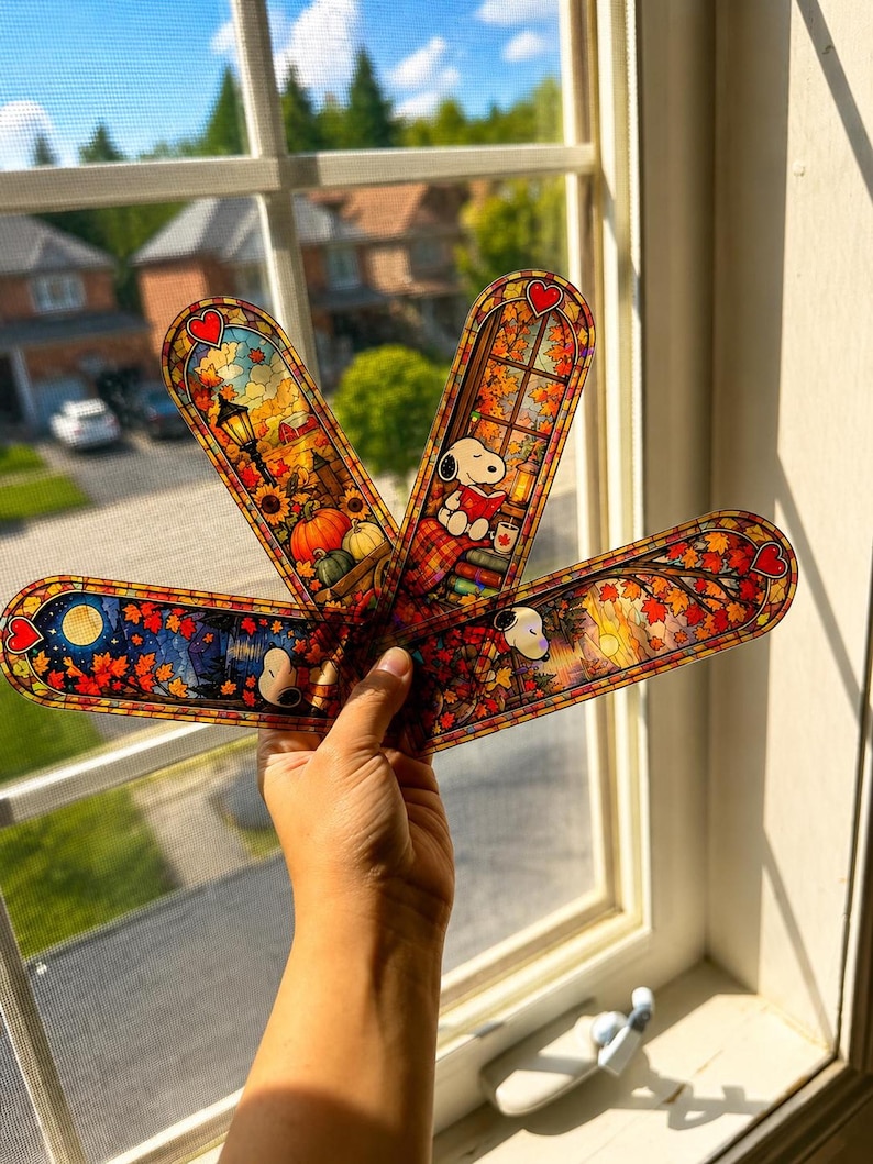 A hand holds up four fan-arranged holographic bookmarks with fall-themed stained glass designs against a window.
