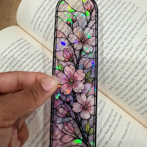 May include: A bookmark with a stained-glass effect, depicting pink cherry blossom flowers and green leaves. The bookmark is framed in black and is held over an open book.
