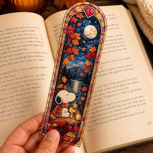 A hand holds a stained-glass bookmark showing Snoopy sitting under a starry night sky with red autumn leaves.