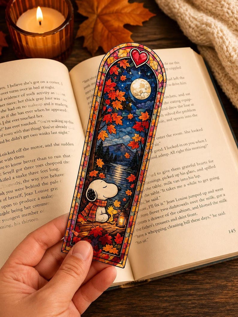 A hand holds a stained-glass bookmark showing Snoopy sitting under a starry night sky with red autumn leaves.