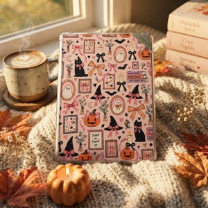 May include: A clear Kindle e-reader case featuring a repeating pattern on a light pink background. The design includes black cats, orange pumpkins, ghosts, witch hats, candles, floral frames, and bows. The text "Trick or Treat" is visible.