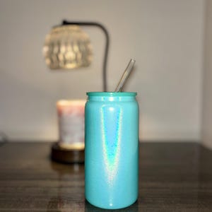 K&ouml;nnte beinhalten: Ein t&uuml;rkis glitzernder Becher mit einem durchsichtigen Strohhalm. Der becherf&ouml;rmige Becher steht auf einer dunklen, reflektierenden Oberfl&auml;che. Im Hintergrund sind eine verschwommene Lampe und eine Kerze zu sehen. Becherh&ouml;he ca. 15 cm.