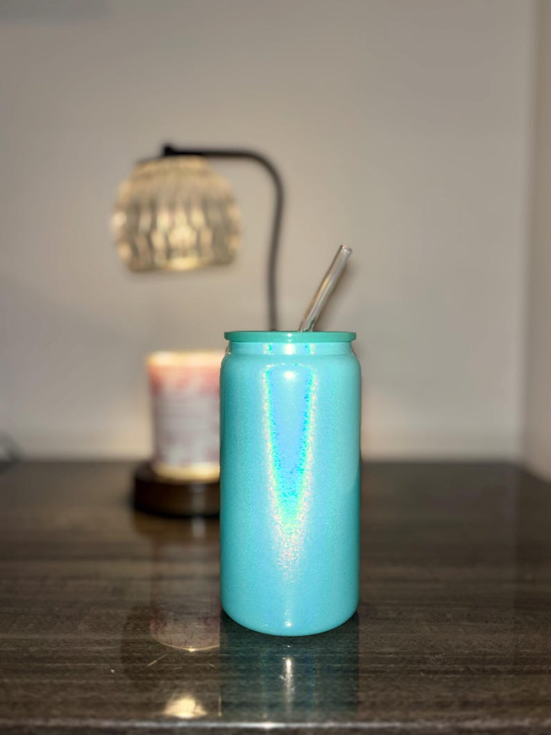 K&ouml;nnte beinhalten: Ein t&uuml;rkis glitzernder Becher mit einem durchsichtigen Strohhalm. Der becherf&ouml;rmige Becher steht auf einer dunklen, reflektierenden Oberfl&auml;che. Im Hintergrund sind eine verschwommene Lampe und eine Kerze zu sehen. Becherh&ouml;he ca. 15 cm.
