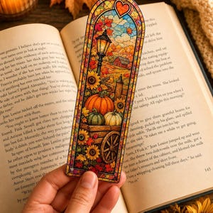 A hand holds an autumn-themed stained-glass bookmark featuring a pumpkin cart on an open book.