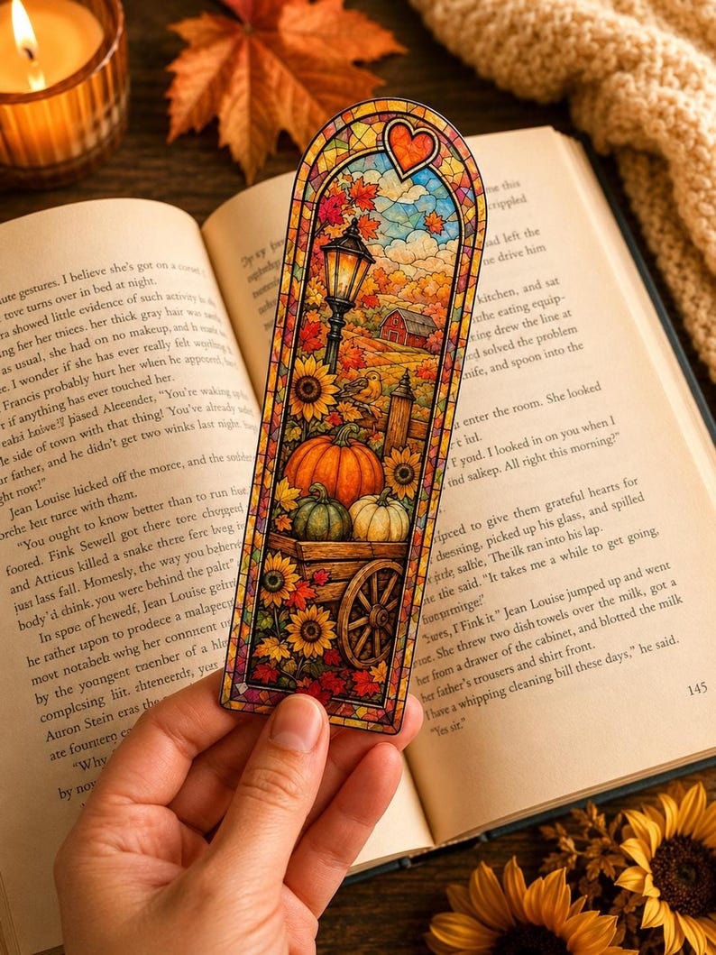 A hand holds an autumn-themed stained-glass bookmark featuring a pumpkin cart on an open book.