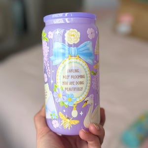 K&ouml;nnte beinhalten: Ein lavendelfarbener Becher mit passendem Deckel. Der Becher ist mit floralen und Insektenillustrationen, einer blauen Schleife und dem Text "Darling, Keep Blooming You Are Doing Beautifully" verziert.