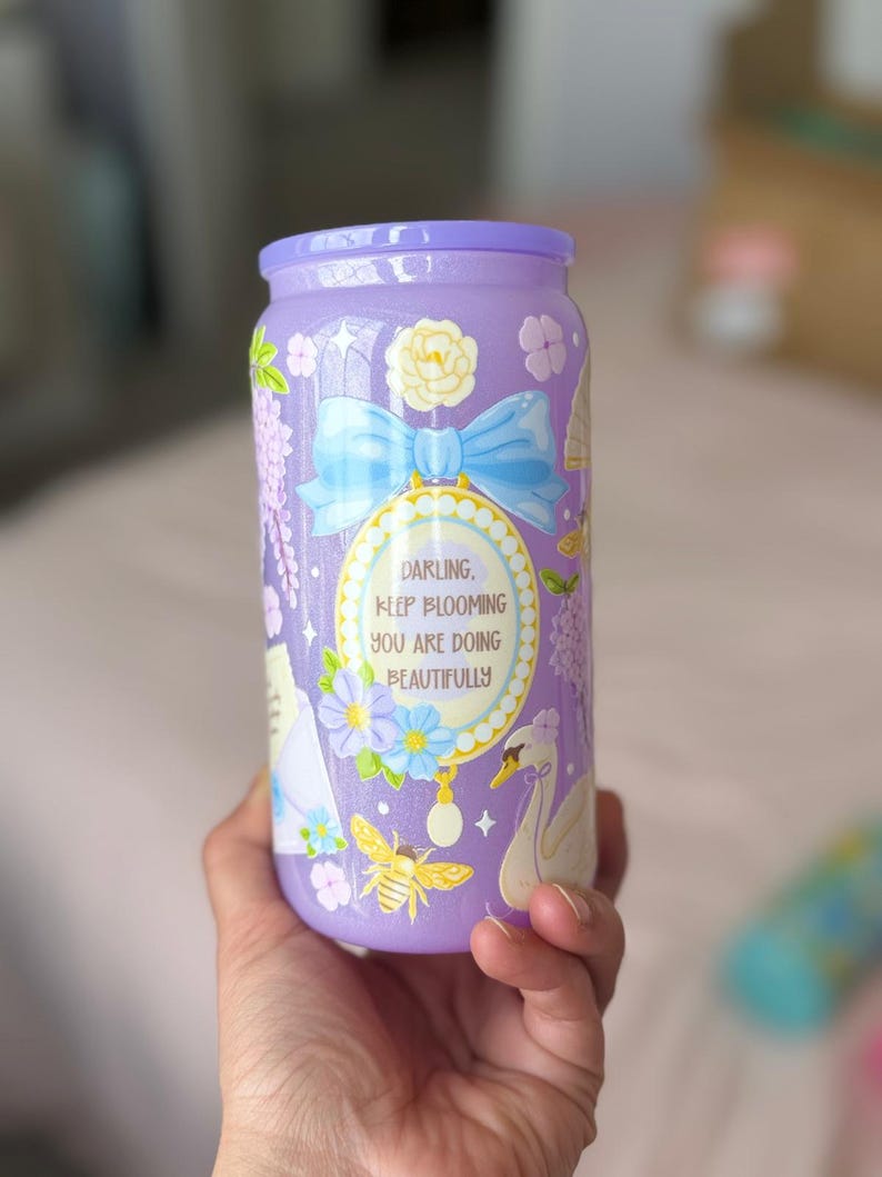 K&ouml;nnte beinhalten: Ein lavendelfarbener Becher mit passendem Deckel. Der Becher ist mit floralen und Insektenillustrationen, einer blauen Schleife und dem Text "Darling, Keep Blooming You Are Doing Beautifully" verziert.