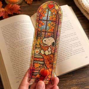 A hand holds a stained-glass bookmark of Snoopy reading by a window with autumn leaves and a pumpkin.