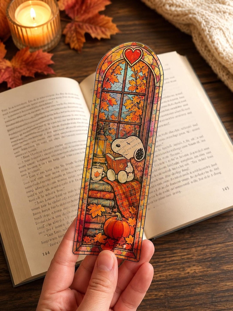 A hand holds a stained-glass bookmark of Snoopy reading by a window with autumn leaves and a pumpkin.