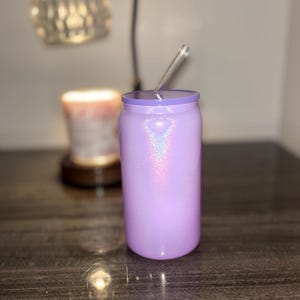 K&ouml;nnte beinhalten: Ein lavendelfarbener Glitzerbecher mit einem durchsichtigen Strohhalm. Der Becher ist zylindrisch mit einem passenden Deckel. Im Hintergrund befinden sich eine Lampe und eine Kerze.