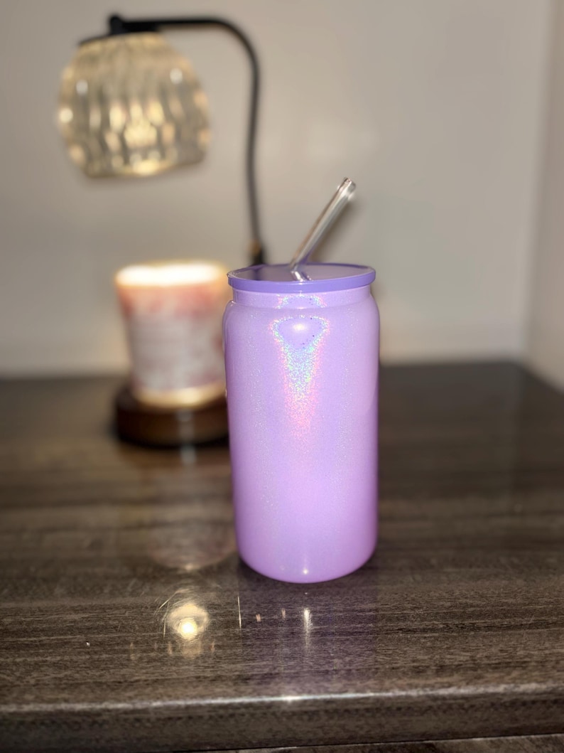 K&ouml;nnte beinhalten: Ein lavendelfarbener Glitzerbecher mit einem durchsichtigen Strohhalm. Der Becher ist zylindrisch mit einem passenden Deckel. Im Hintergrund befinden sich eine Lampe und eine Kerze.