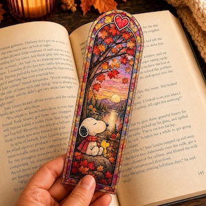 A hand holds a stained-glass bookmark featuring Snoopy reading under an autumn tree at sunset.