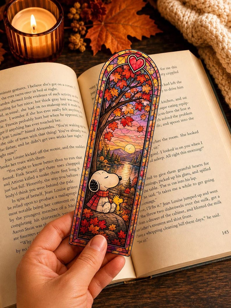 A hand holds a stained-glass bookmark featuring Snoopy reading under an autumn tree at sunset.