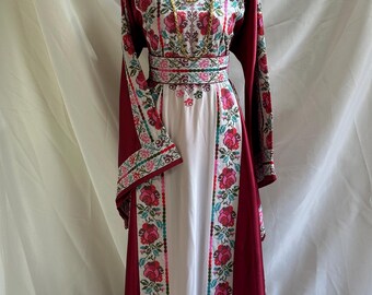 May include: A long, embroidered dress with a white bodice and burgundy sleeves and side panels. The dress features floral embroidery in shades of red, pink, and green. A gold necklace is draped around the neckline. The dress is displayed on a mannequin.