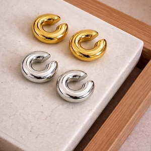 Puede incluir: Cuatro puños de oreja, dos dorados y dos plateados, se exhiben sobre una superficie rectangular de color claro. Los puños de oreja tienen un diseño liso y redondeado, con forma de C. El fondo es una bandeja de madera.