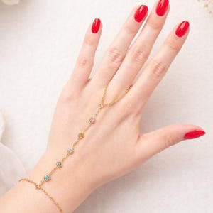 Puede incluir: Una pulsera de cadena de mano dorada con piedras preciosas de colores. La pulsera se conecta a un anillo y presenta una serie de pequeñas piedras preciosas redondas de varios colores. La mano de la imagen tiene esmalte de uñas rojo.