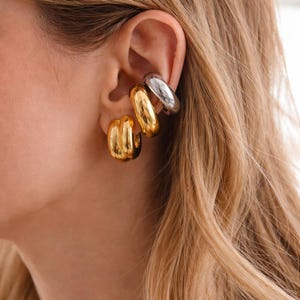 Puede incluir: Primer plano de tres puños de oreja en tonos dorados y plateados. Los puños de oreja se llevan en la oreja. Los puños de oreja dorados tienen un diseño acanalado. El puño de oreja plateado es liso y brillante. La persona tiene el pelo rubio.