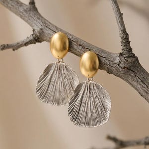 Puede incluir: Un par de pendientes con una parte superior dorada de forma ovalada y una parte inferior plateada en forma de abanico. Los pendientes se muestran en una rama marrón sobre un fondo borroso. Los pendientes tienen un diseño texturizado y acanalado.