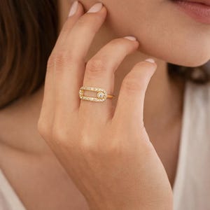 Puede incluir: Un anillo de oro con un diseño rectangular, incrustado con pequeñas piedras preciosas transparentes. Una piedra preciosa más grande está engastada en el centro del rectángulo. El anillo se lleva en un dedo, con la mano y parte de una cara visibles.