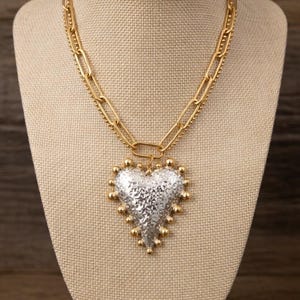 Puede incluir: Un collar dorado con un gran colgante en forma de corazón plateado texturizado. El corazón está bordeado con pequeñas cuentas doradas. El collar se exhibe sobre una cabeza de maniquí beige sobre un fondo de madera.