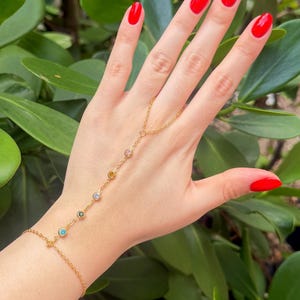 Puede incluir: Una pulsera de cadena de mano dorada con piedras preciosas de colores. La pulsera conecta desde la muñeca hasta el dedo medio. Las piedras preciosas son en tonos de azul, verde, amarillo, rosa y morado. Las uñas están pintadas de rojo.