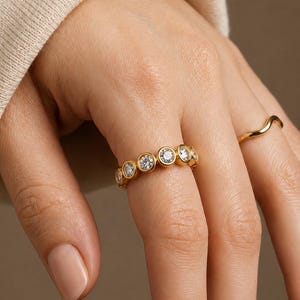 Puede incluir: Un anillo de oro con una serie de piedras preciosas redondas y transparentes engastadas en un diseño de bisel. El anillo está en un dedo, y también es visible otro anillo de oro con un diseño curvo.