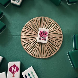 May include: A white Mahjong tile featuring a red graphic of a stylized dragon face and the text CRAK, centered on a circular gold-colored decorative mat with a radiating sunburst pattern, surrounded by various green and white Mahjong game tiles.