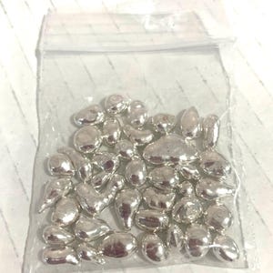 May include: A transparent plastic bag containing many small, irregularly shaped, shiny silver metal pieces. The pieces have a smooth, reflective surface, and vary in size and form, resembling droplets or pebbles. The bag is partially visible, showing the contents within.