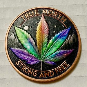 May include: A circular, copper-toned coin featuring a vibrant cannabis leaf design against a black backdrop. The leaf is rendered in hues of purple, green, yellow, and red. The coin is inscribed with the words "TRUE NORTH" and "STRONG AND FREE".