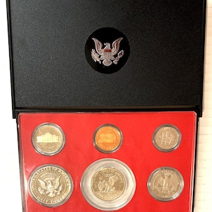 Op de afbeelding: Een zwarte presentatiekoffer met een rode binnenkant bevat een set Amerikaanse munten. De munten omvatten een halve dollar, een dollar, een penny, een nickel, een dime en een kwart dollar. De koffer heeft een cirkelvormig embleem.