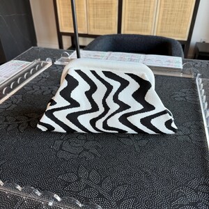May include: A structured clutch bag featuring a bold black and white wavy, zigzag pattern. The bag has a curved top frame and a textured, knit-like fabric construction. The design is symmetrical, with the black and white lines creating a continuous, flowing visual effect across the entire surface of the accessory.