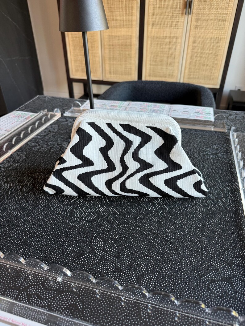 May include: A structured clutch bag featuring a bold black and white wavy, zigzag pattern. The bag has a curved top frame and a textured, knit-like fabric construction. The design is symmetrical, with the black and white lines creating a continuous, flowing visual effect across the entire surface of the accessory.