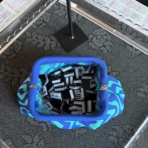 May include: A blue and teal patterned fabric pouch with a metal frame closure, filled with a collection of small, rectangular black and white Mahjong tiles featuring various engraved symbols and characters in green, red, and blue ink.